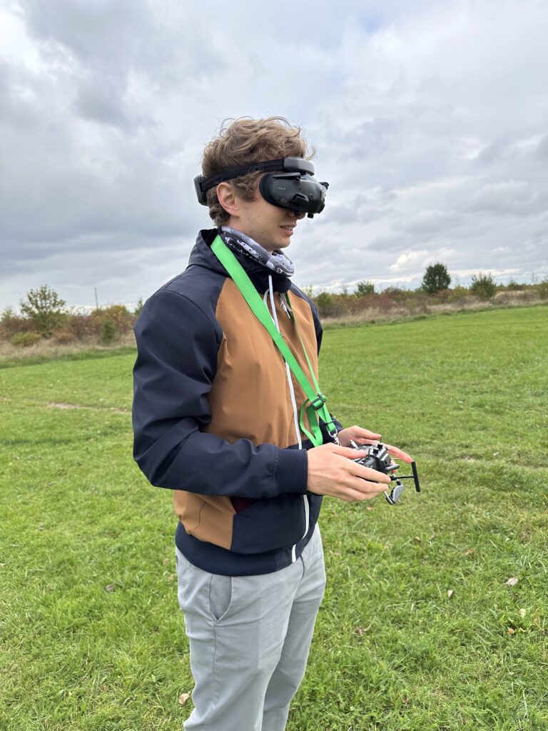 Osoba, která nosí FPV brýle a drží modelářskou vysílačku dronu, stojí na travnaté louce pod zamračenou oblohou.
