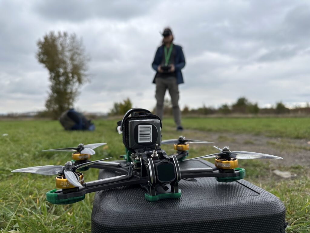 Dron s namontovanou kamerou a čtyřmi vrtulemi spočívá na černém pouzdře na travnatém poli. V pozadí osoba, která drží dálkové ovládání v pozadí pod oblačnou oblohou.