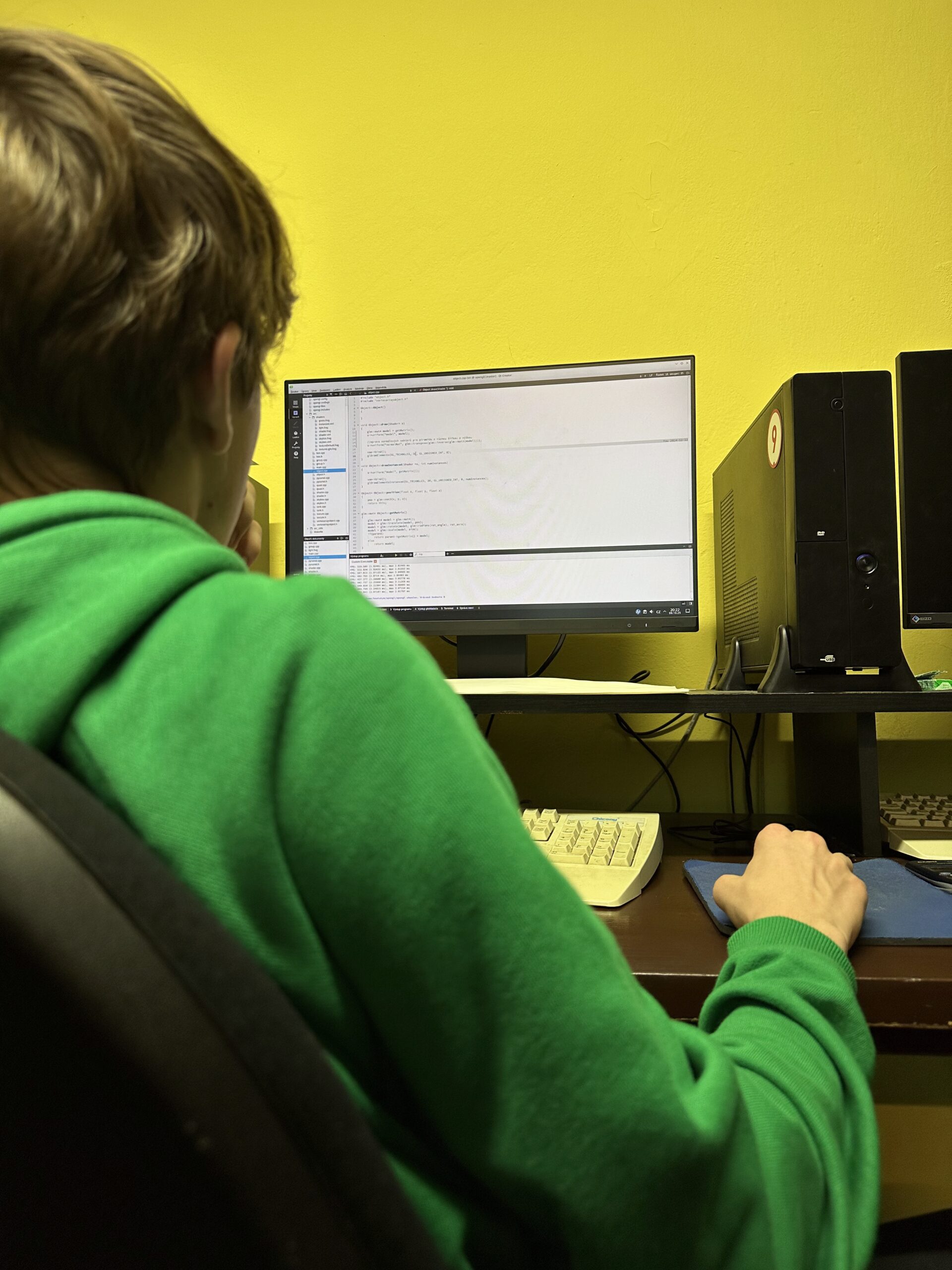Student v zelené mikině pomocí počítače s monitorem zobrazující text, žlutá stěna pozadí.