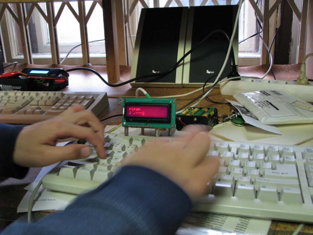 Ruce píšící na klávesnici se zeleným elektronickým modulem s nápisem "pípám J .---" a počítačovými reproduktory v blízkosti okna.
