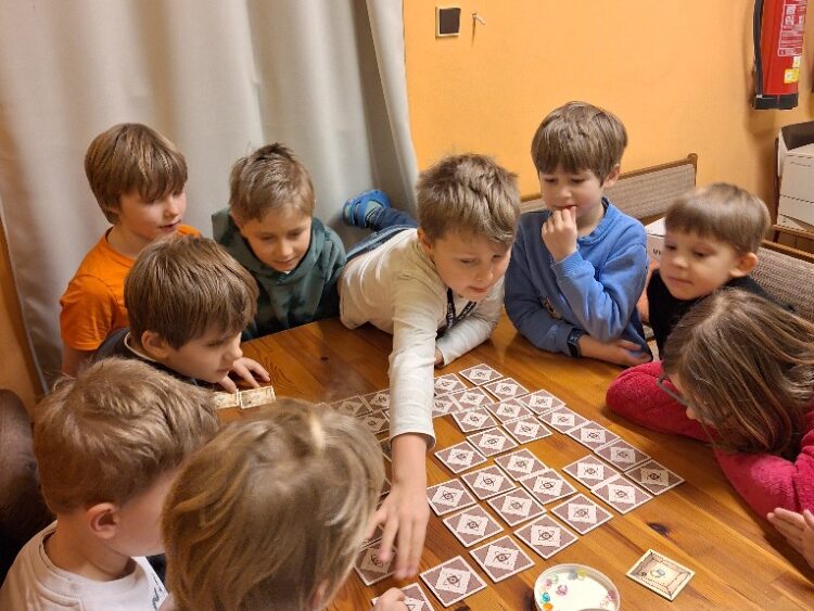 Skupina dětí se shromáždila kolem dřevěného stolu a hrála deskovou hru s dlaždicemi a krabicí plnou barevných předmětů.