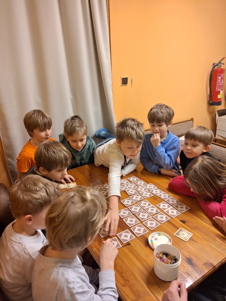 Skupina dětí se shromáždila kolem dřevěného stolu a hrála deskovou hru s dlaždicemi a krabicí plnou barevných předmětů.