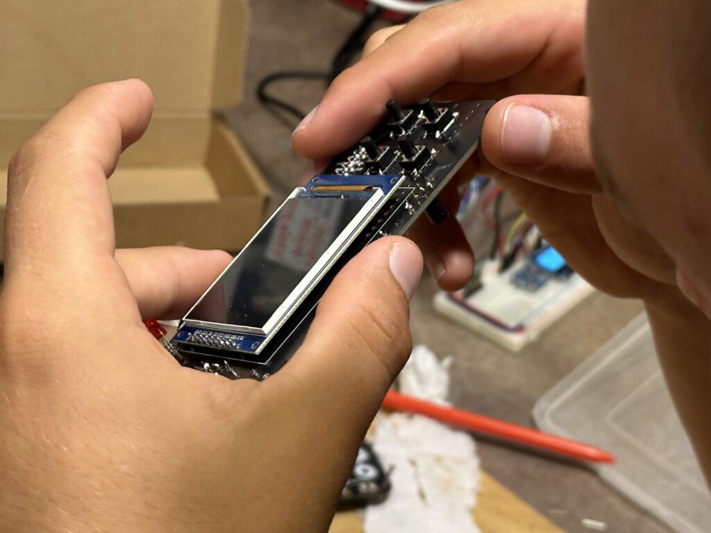 Dvě ruce drží plošný spoj s displejem a elektronickými součástkami, lepenková krabička v pozadí na stole, dráty, štětec.