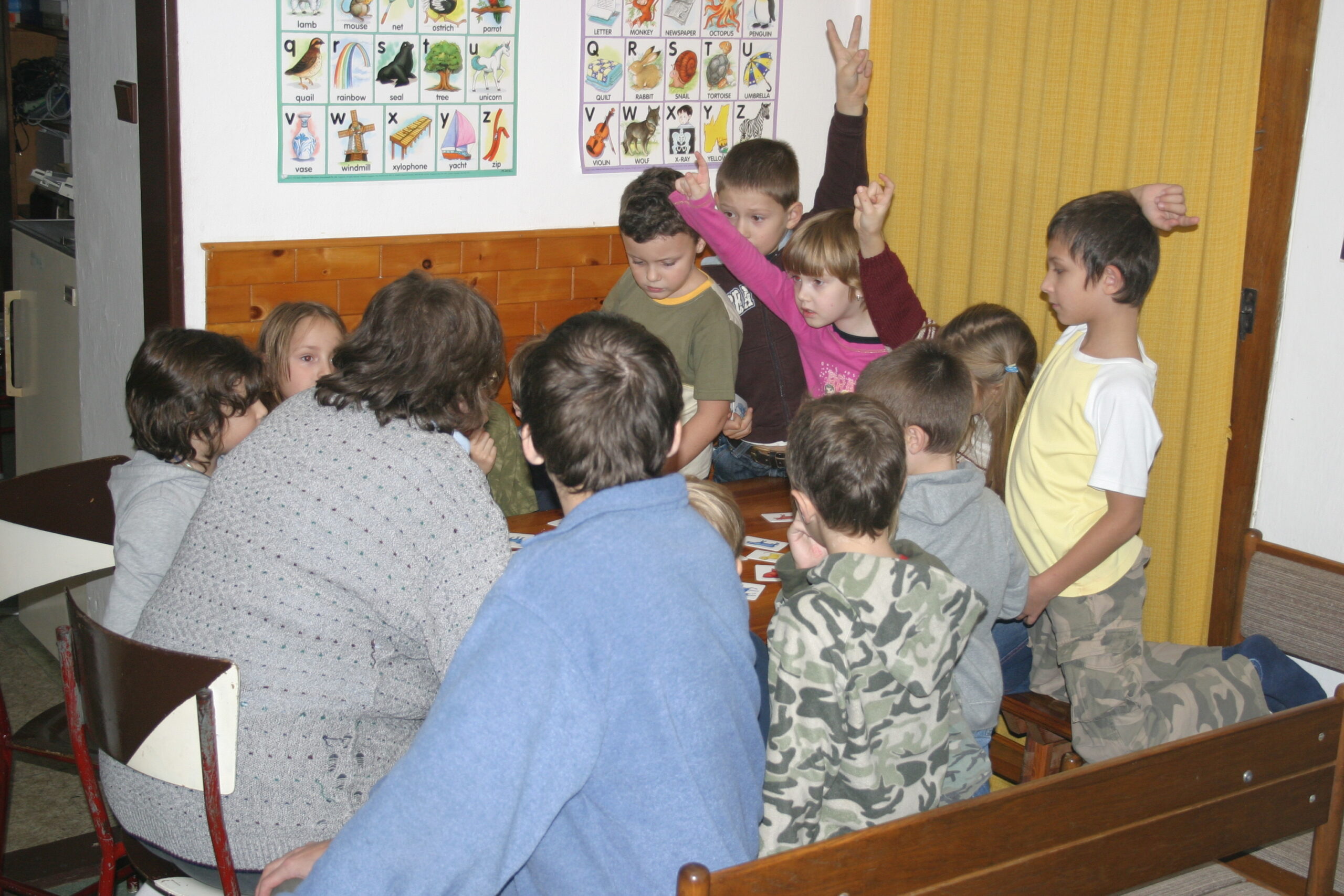 Děti se shromáždily kolem stolu v místnosti s nástěnnými vzdělávacími tabulkami, některé zvedly ruce.