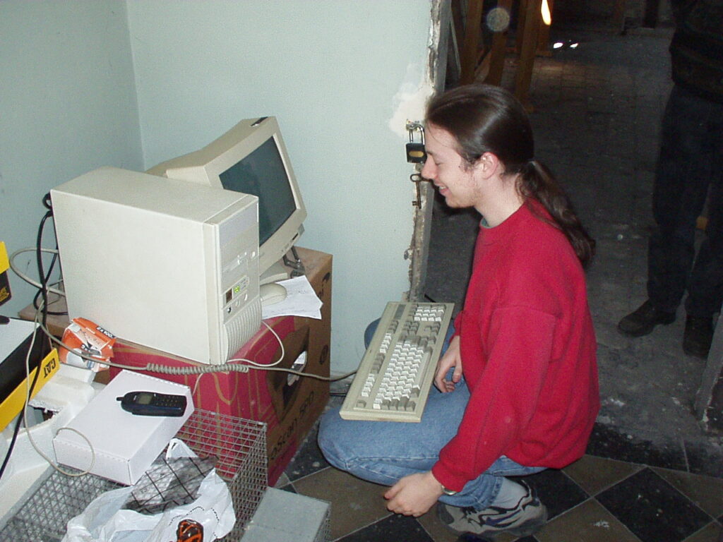 Osoba v červené mikině drží klávesnici vedle starého béžového PC a monitoru na papírové krabici, s mobilním telefonem na nedaleké bílé krabičce.