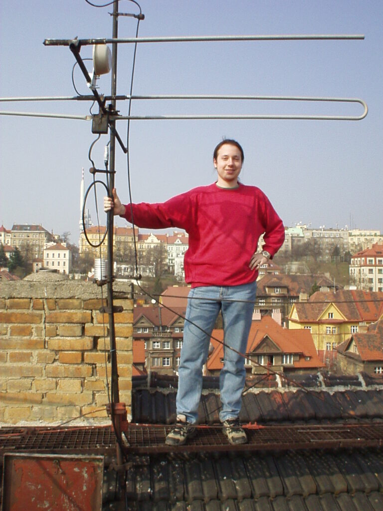 Osoba v červené mikině stojí na střeše drží se antény, s výhledem na městké budovy s červenými střechami v pozadí.