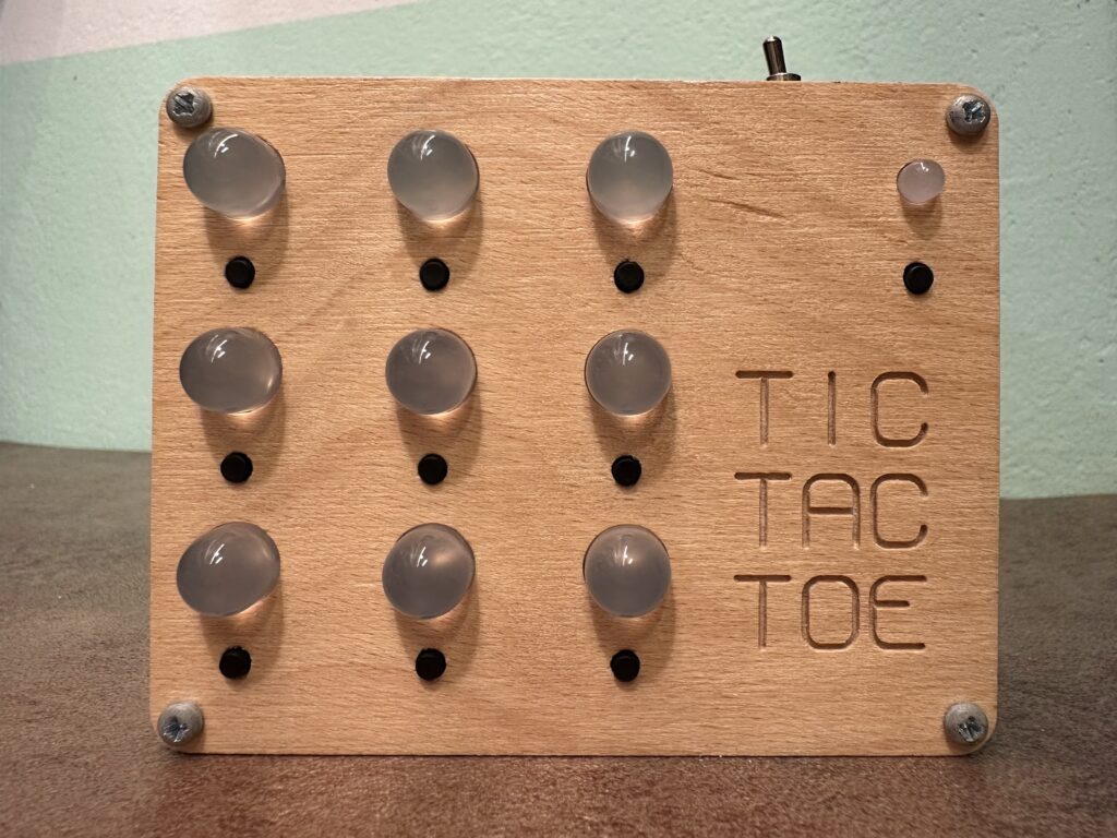 Elektronická hra „Tic Tac Toe" s dřevěným panelem, velkými LED svítivkami, černými tlačítky a vyfrézovaným nápisem.