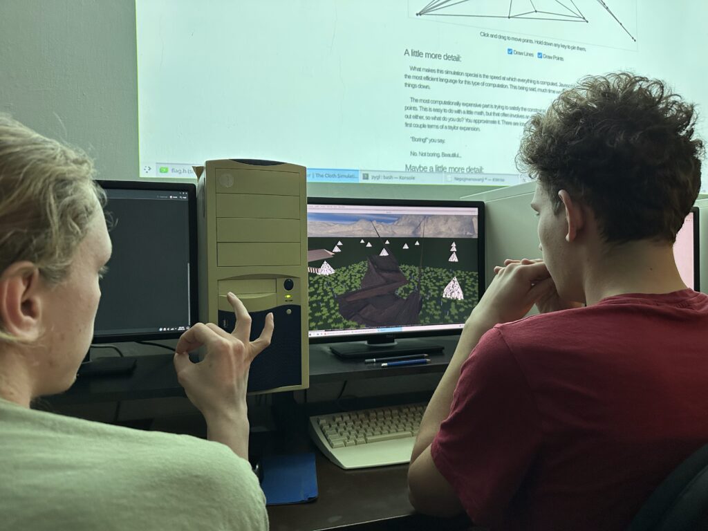 Dva studenti u počítačů, jeden ukazuje ou kej, druhý sleduje monitor s 3D simulací, nad hlavou projektor s diagramy.