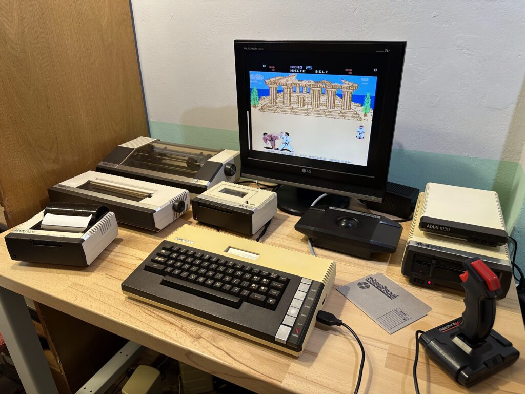 Retro počítačová sestava Atari 800 XL s množstvím přislušenství, na monotoru se ukazuje bojová hra a joystick.