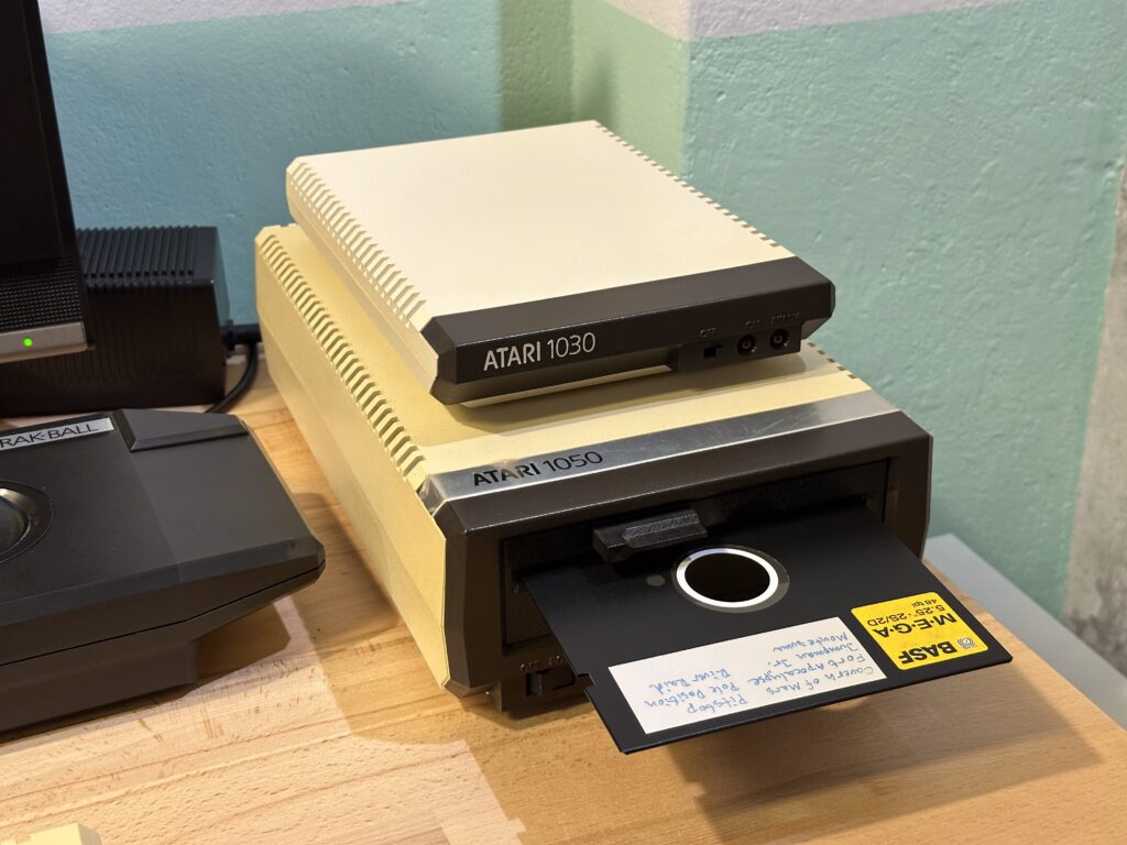 Dvě retro zařízení Atari: disketová mechanika Atari 1050 s popsanou disketou a Atari 1030 na dřevěném stole.