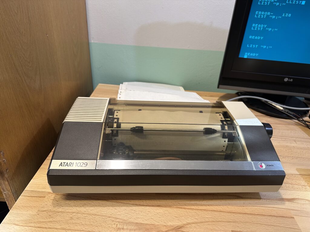 Tiskárna Atari 1029 na dřevěném stole s LG monitorem zobrazujícím modrý text obrazovky.